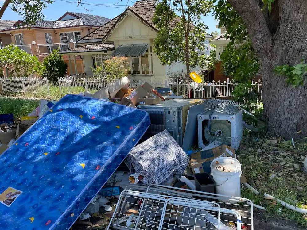 Aussie street with hard rubbish collection: old mattress and junk piled up near a house under a sunny sky.