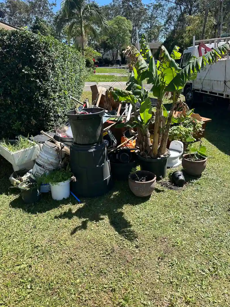 Messy backyard with green waste removal: old pots, banana plants, and junk piled up beside a white ute under the Aussie sun.