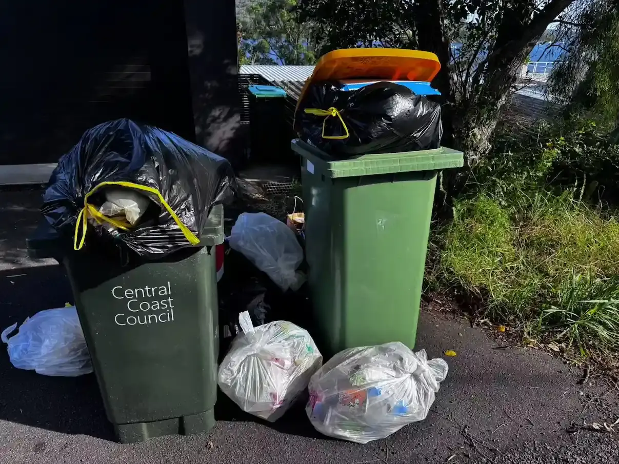 Aussie waste collection service: Central Coast Council bins overflowing with black and white bags on a quiet street.