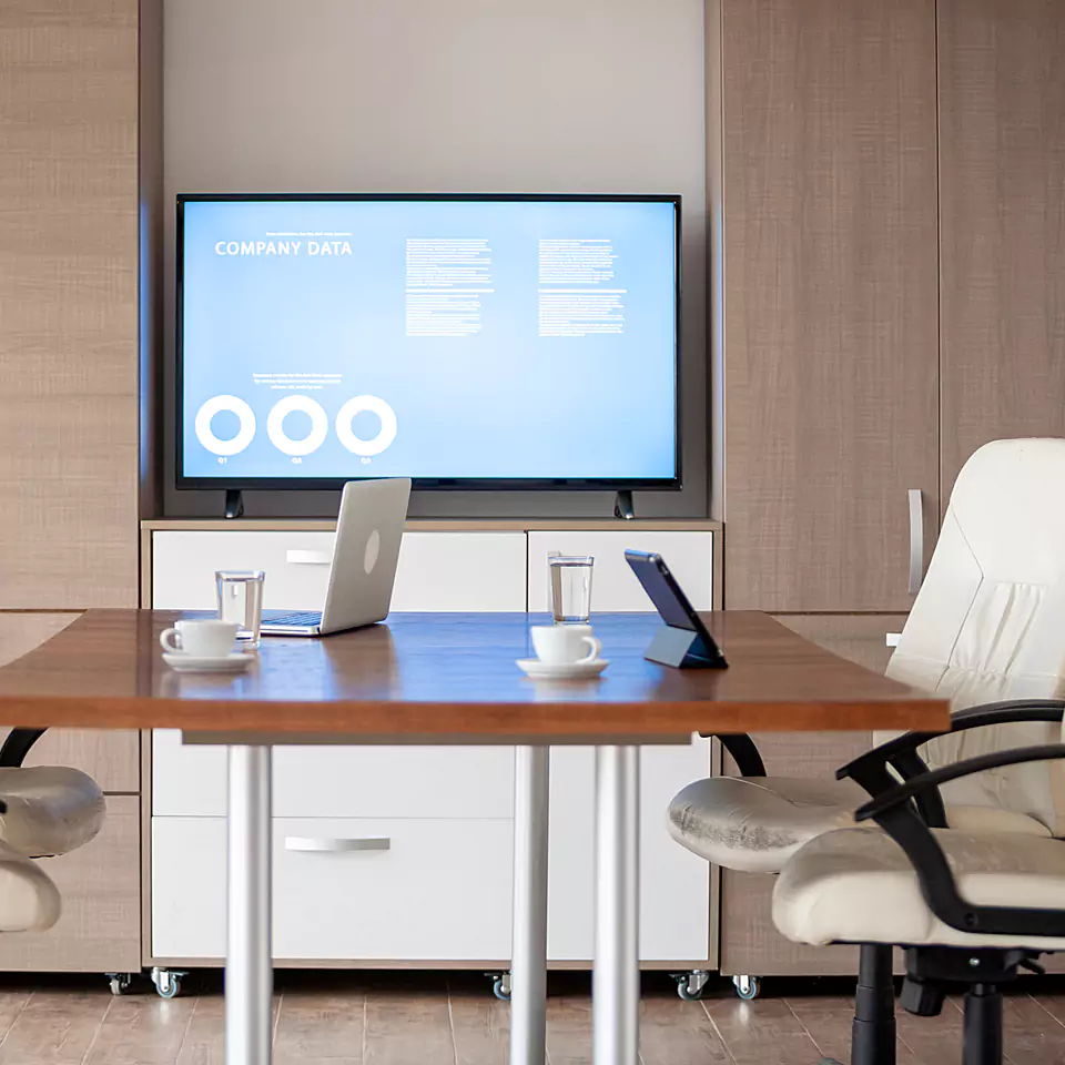 Commercial office AV equipment including a large presentation display, laptop, and tablet in a corporate meeting room setup.